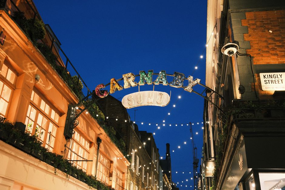 A vibrant street scene in Soho featuring a colorful, illuminated sign that reads 'CARNABY', with each letter in a different bright color, mounted on a metal arch spanning the narrow pedestrian street.