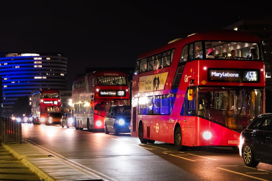 A row of three red double-decker buses and several parked cars on a city street at night. The buses are illuminated by interior and exterior lights, with visible route displays reading 'Oxford Circus'