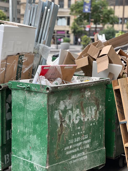 A large, weathered green waste skip filled with various types of rubbish, including flattened cardboard boxes, partially torn and crumpled packaging, and miscellaneous debris. The skip is positioned o